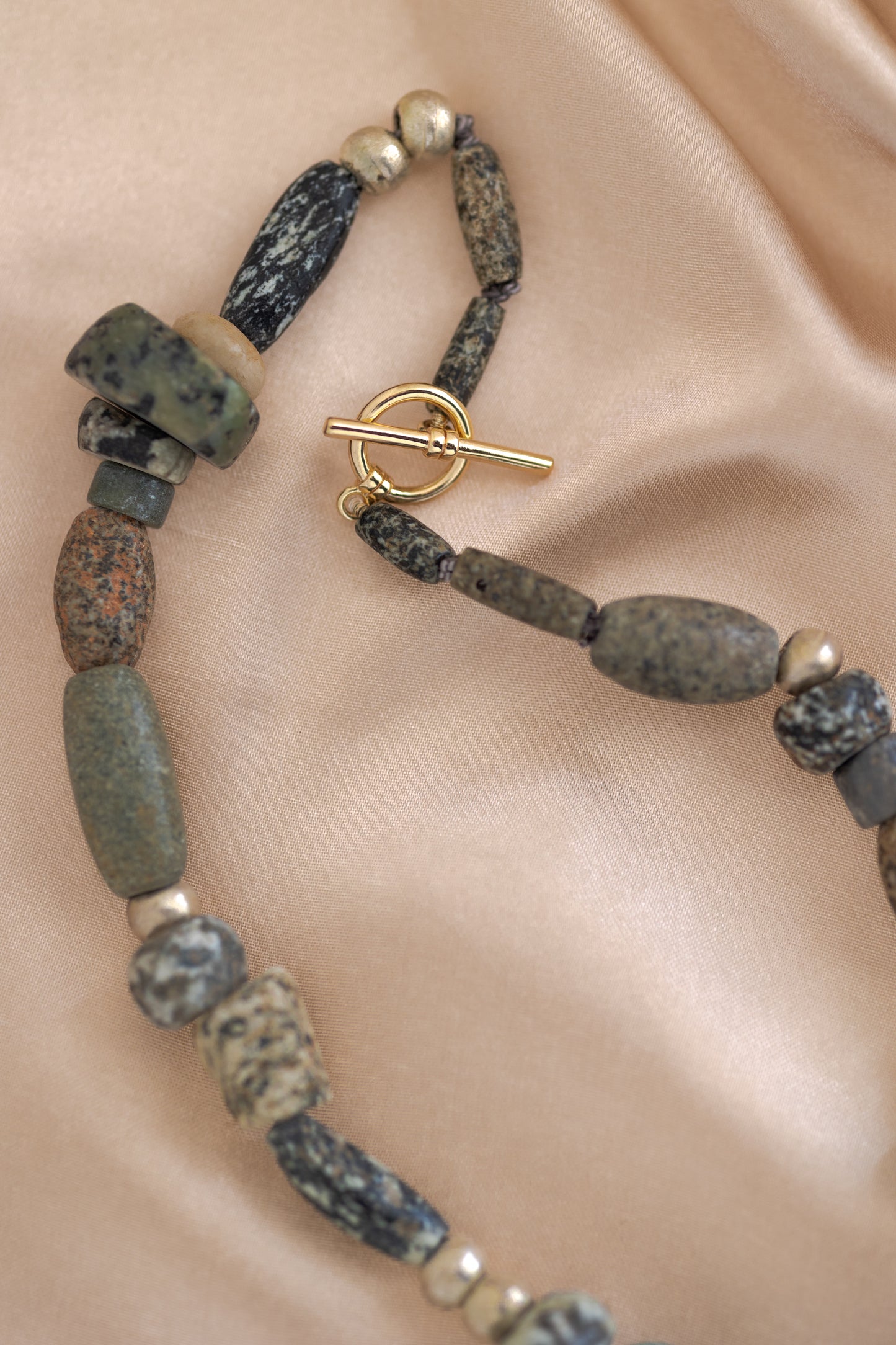 Close up of an ancient mali granite, ancient mali quartz, ethiopian metal and turquoise beaded necklace with a gold toggle clasp on a silky beige background