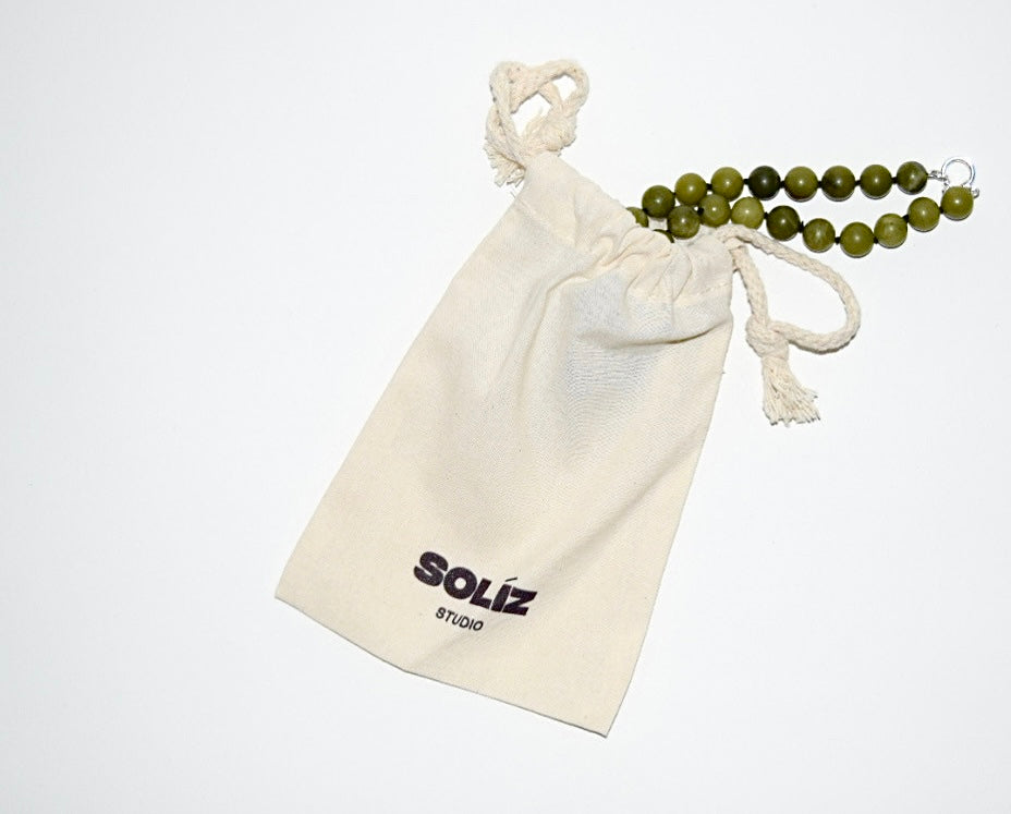 12mm chinese jade beaded necklace coming out of a cream colored cotton drawstring bag that has a studio soliz logo in black on the bottom front of the bag.