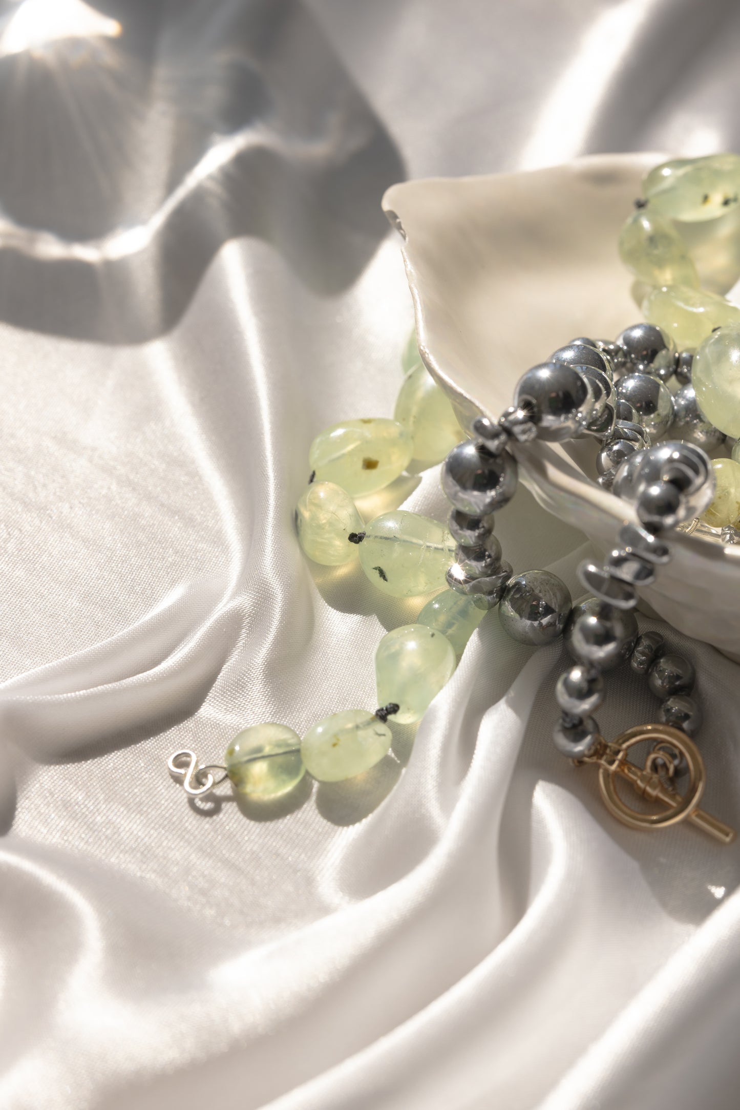 Close up image of a hematite beaded necklace and perinite beaded necklace in a bowl on a silky, shiny cloth material